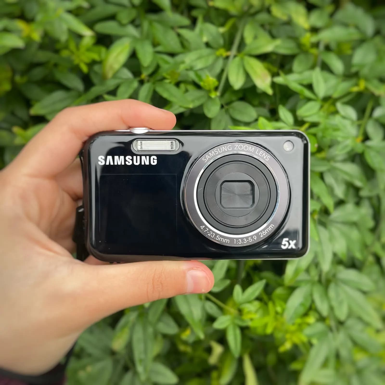 Cámara Samsung negra brillante sostenida en una mano, de frente, con el objetivo de 5 aumentos